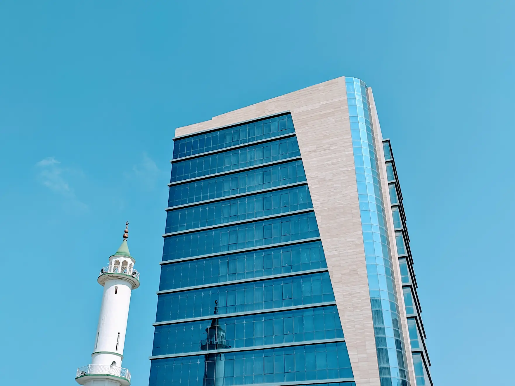 A glass office tower beside a white minaret under a clear blue sky.